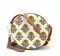 Round crossbody bag with cream and mustard block print floral pattern, tan leather trim and gold tassel charm